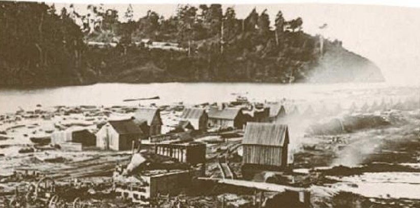 Early Northwest logging settlement — mill smoke, cabins, and log boom on the water