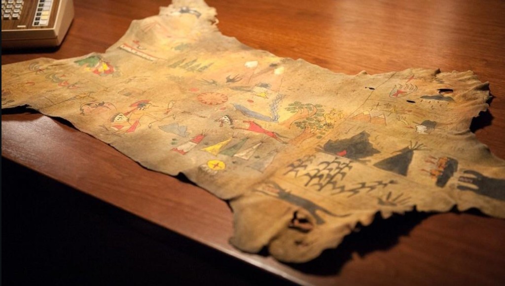 Nez Perce hide map unrolled on a wooden table — painted tipis, peaks, corn, trees, and figures