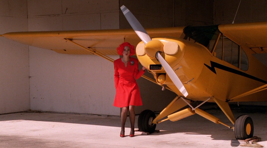 Lil in a red dress beside a yellow airplane - Gordon Cole's Blue Rose briefing